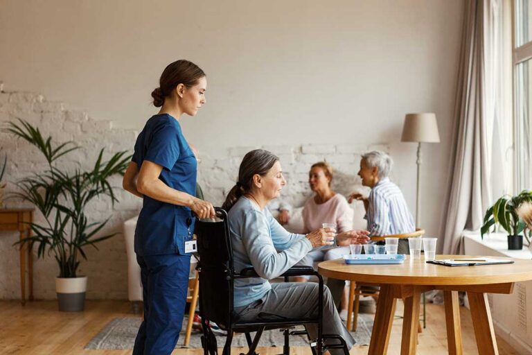 An older adult sits comfortably in a sunlit common area, sipping tea and chatting with another resident nearby.
