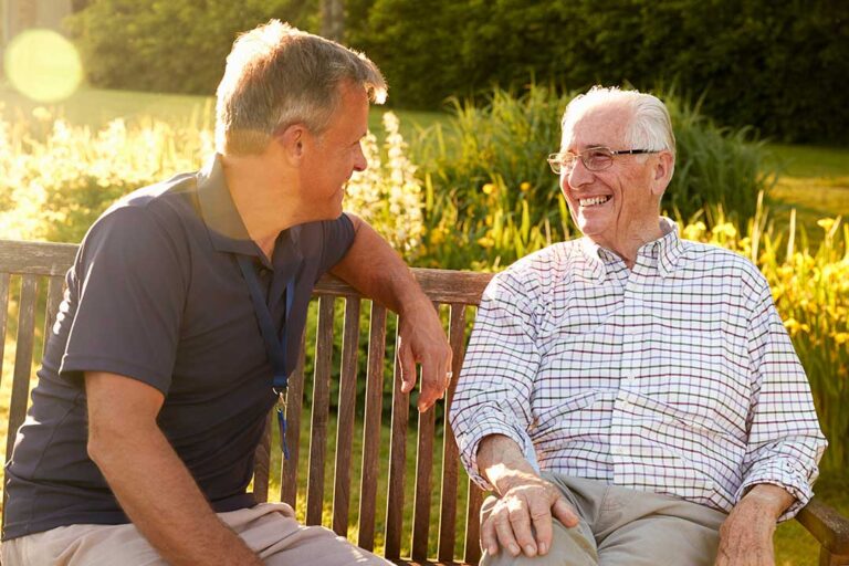 A group of older adults sitting together at a table, talking and laughing in a bright communal living area.
