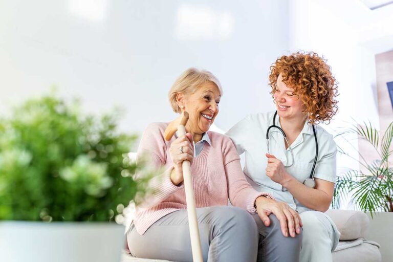 Caregiver calmly helps an older adult with a daily activity in a bright assisted living room.